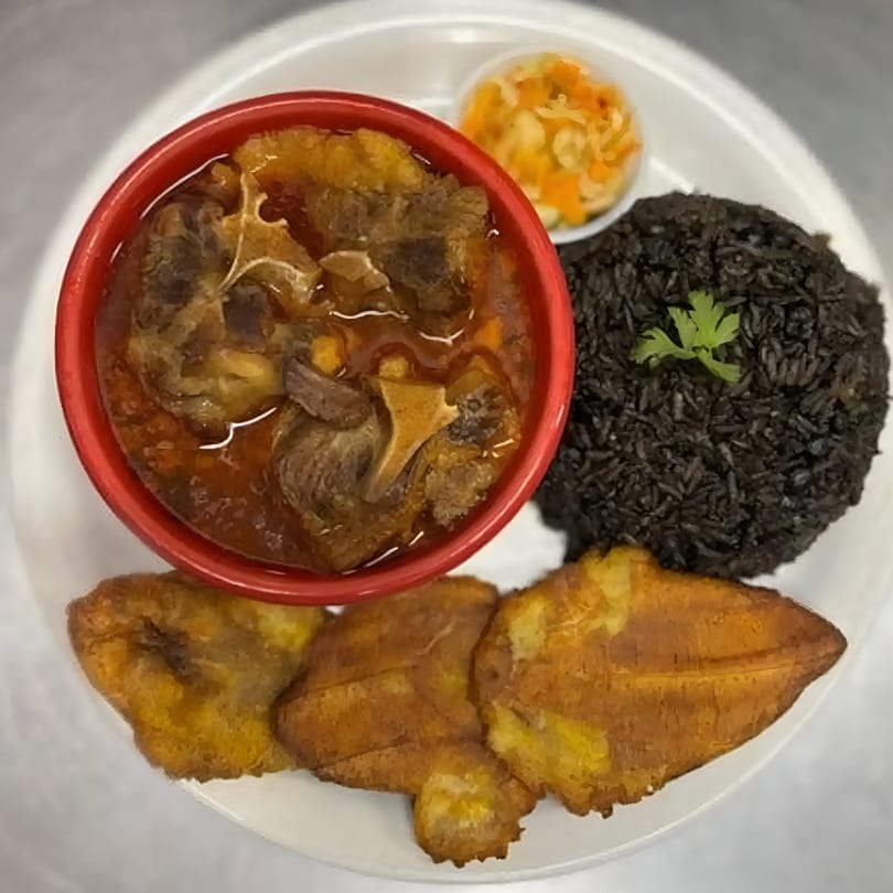 Haitian oxtail stew in a red bowl, black rice, fried plantains, and pikliz on a white plate.
