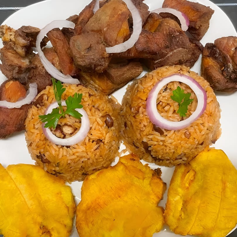Haitian fried pork, rice and beans, and fried plantains garnished with onions and parsley.