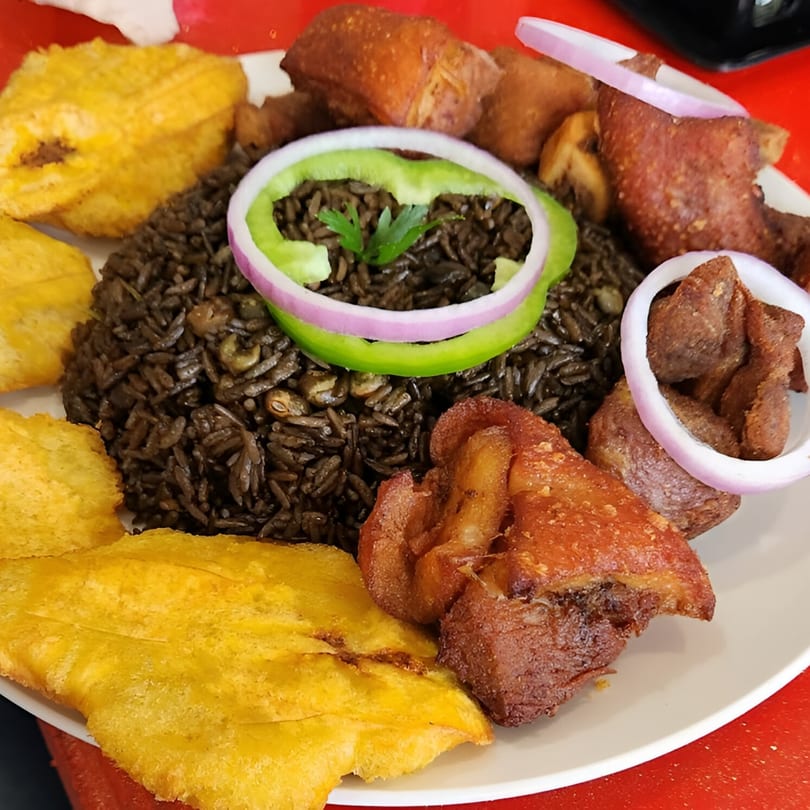 Haitian griot with black rice, fried plantains, and red onion and green pepper garnish.