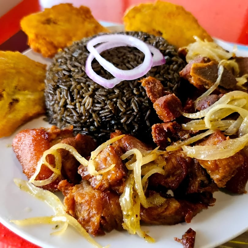 Haitian griot with black rice, fried plantains, and pickled onions on a white plate.