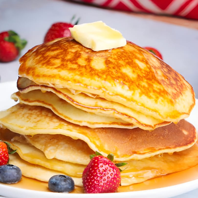 Stack of golden pancakes with butter, syrup, strawberries, and blueberries on a white plate.