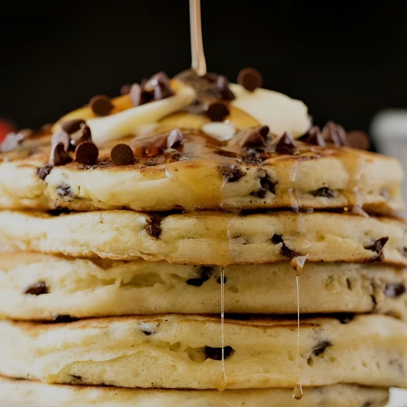 Syrup pouring over a stack of chocolate chip pancakes topped with banana slices.