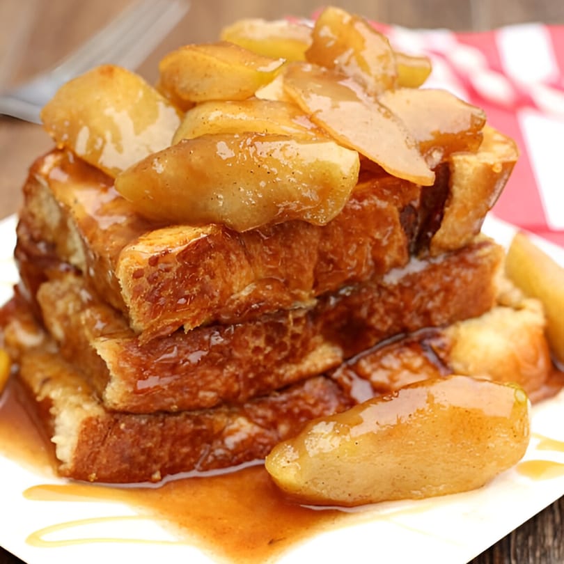 A stack of golden brown Haitian French toast with caramelized apples and syrup on a white plate.