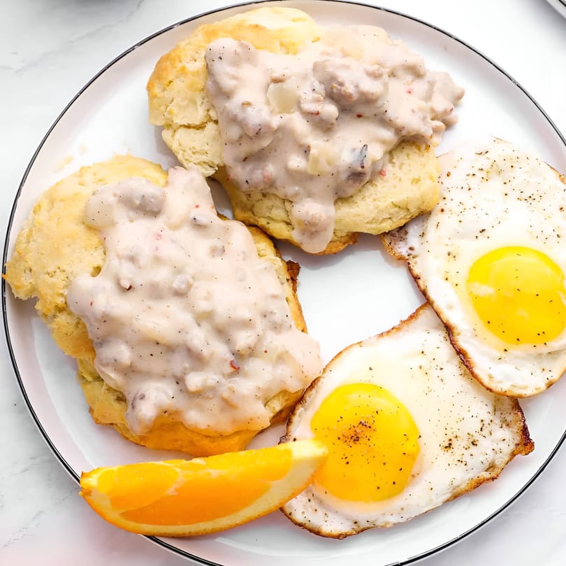 Biscuits and gravy with two fried eggs and an orange slice on a white plate.